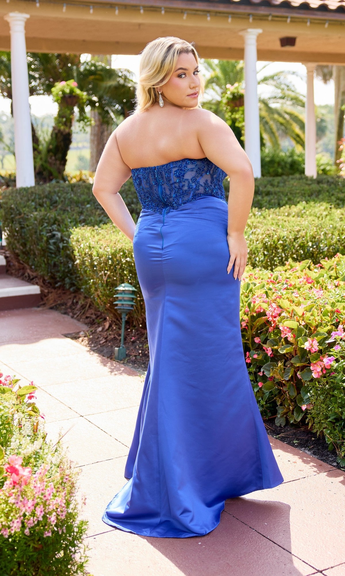 A woman in the Primavera 14109 long plus-size prom dress, featuring a strapless sheer embroidered bodice and satin sheath skirt, stands on a garden path outdoors, glancing over her shoulder.
